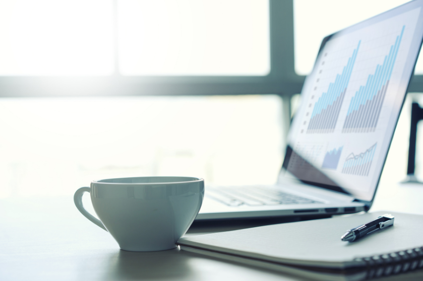 Computer with data on screen sits on a desk with coffee.