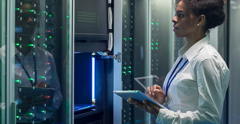 A woman stands in a server room, holding a tablet and taking notes.