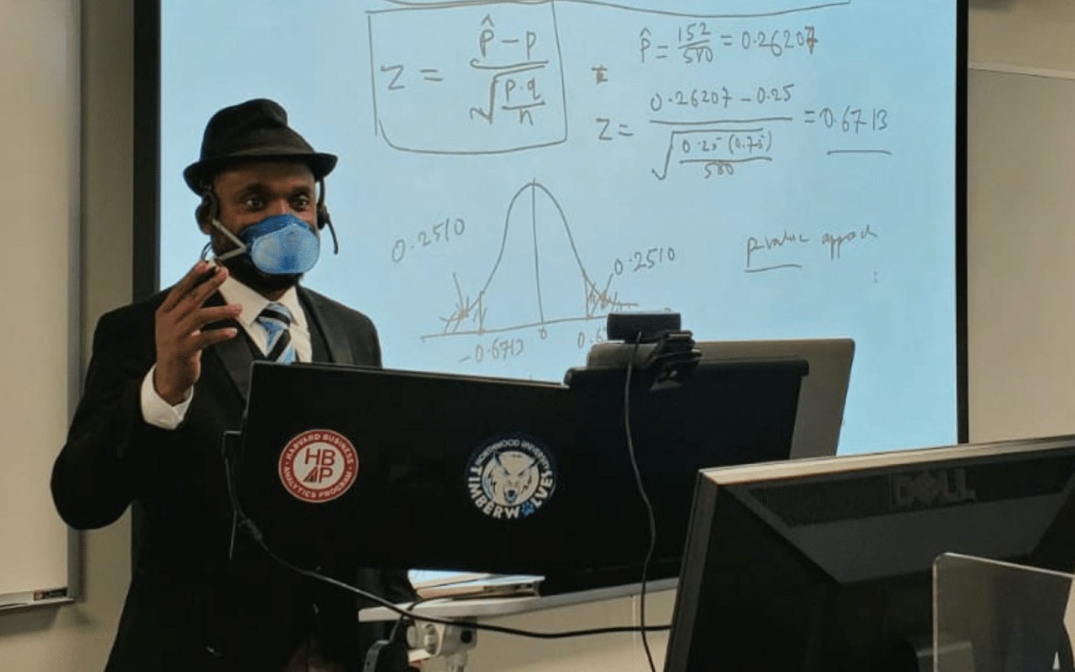 Dr. Itauma wears a mask and stands in front of a projector, teaching a class.